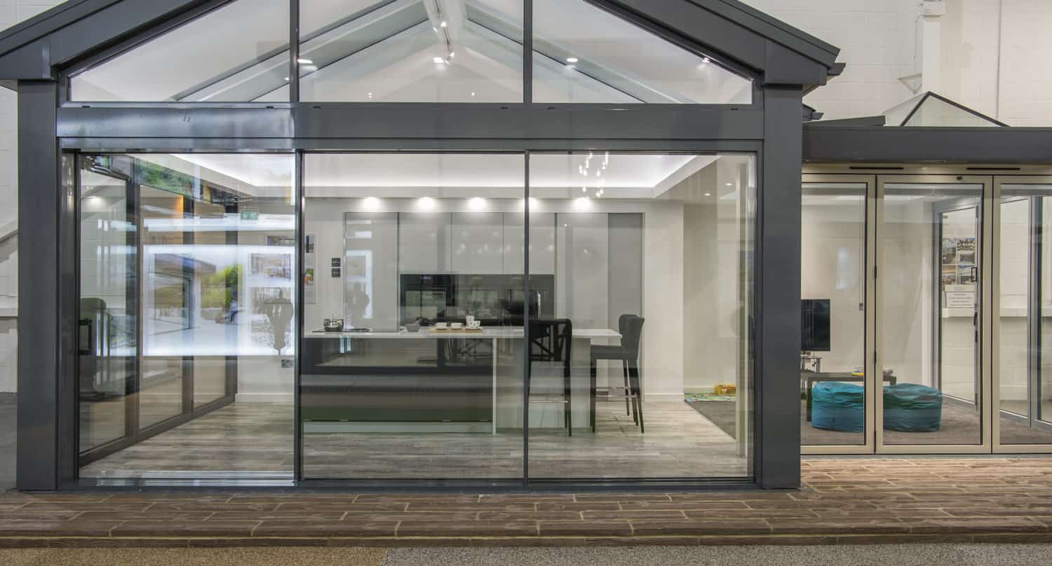 Modern glass-walled room with a peaked roof, featuring a dining table and chairs inside, seen through large bifold doors. The space is bright with contemporary decor, and adjacent to another open area with blue bean bags.