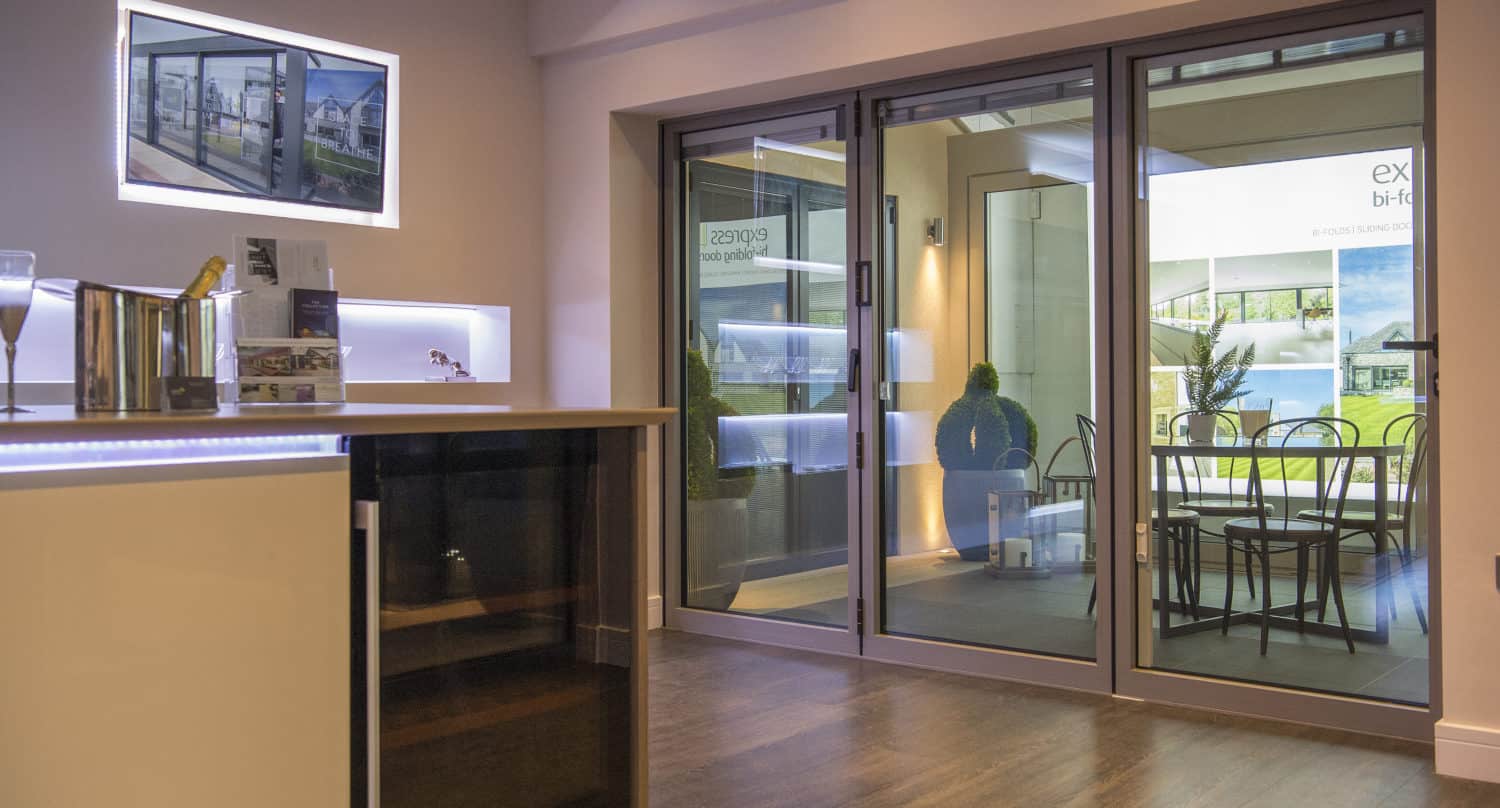 Modern interior showroom featuring bespoke glass solutions, with sliding glass doors opening to a patio area with a table and chairs. The room offers wood flooring, a counter with mini fridge, and wall displays with brochures and illuminated shelves.