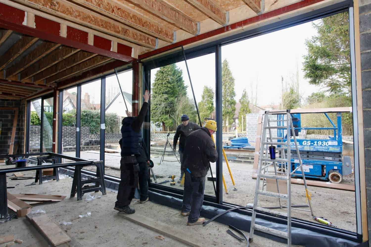 Four construction workers install large glass panels, possibly for sliding doors, in a building under construction, with tools, ladders, and equipment scattered around and an outdoor area visible through the open wall.