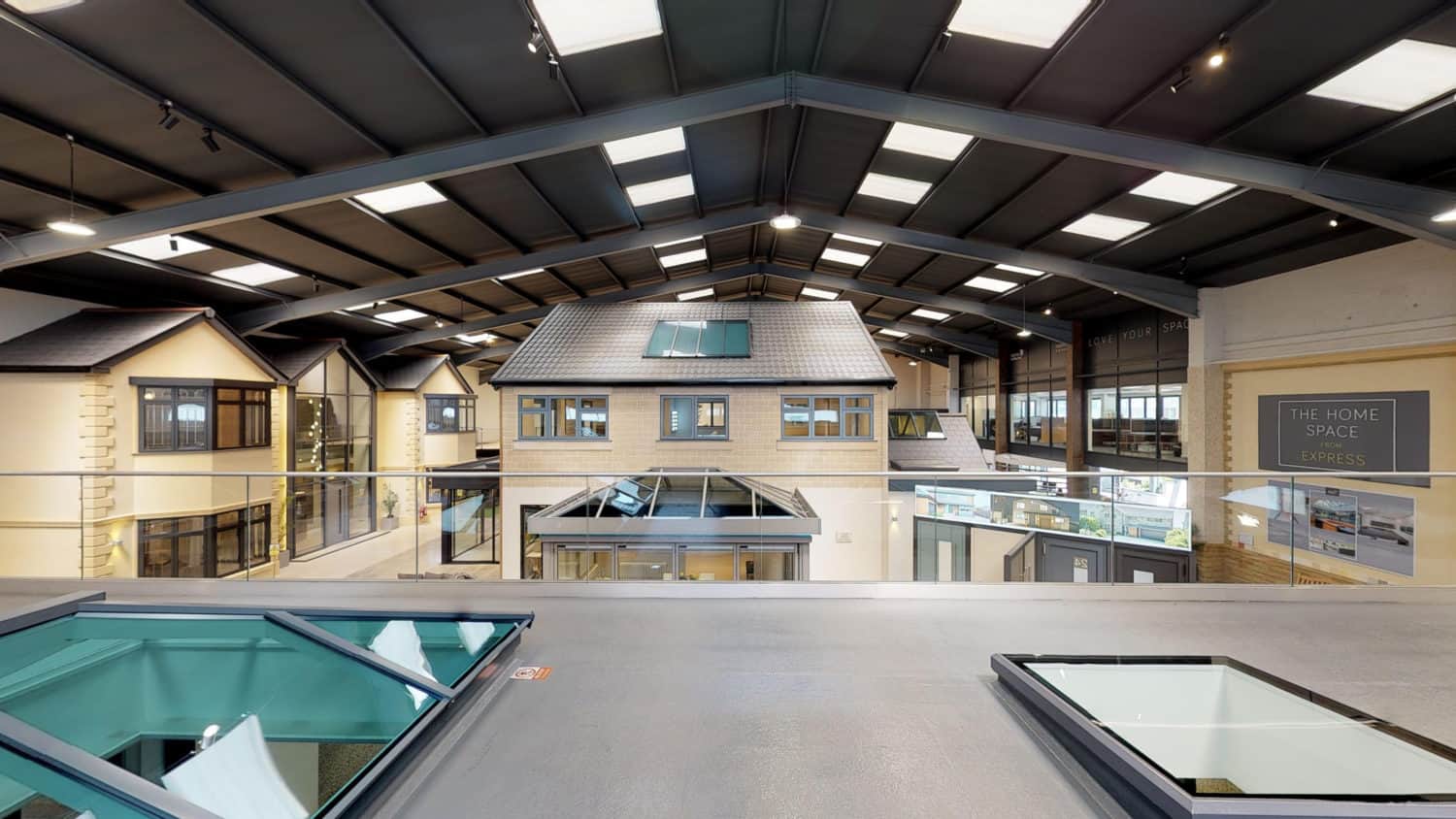 Interior view of a large showroom with high ceilings, skylights, and model house facades. The space features modern lighting, glass railings, bespoke glass solutions, and displays related to home improvement or construction.