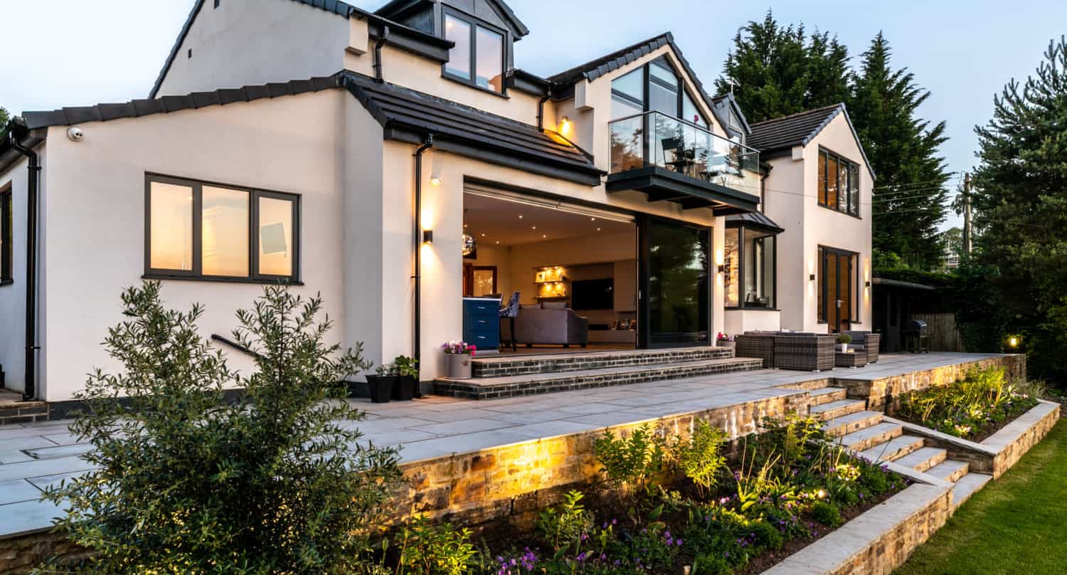Modern two-story house with large windows, sliding doors, and an open patio. A landscaped garden with stone steps and flower beds surrounds the home, while warm indoor lights create a welcoming atmosphere at dusk.
