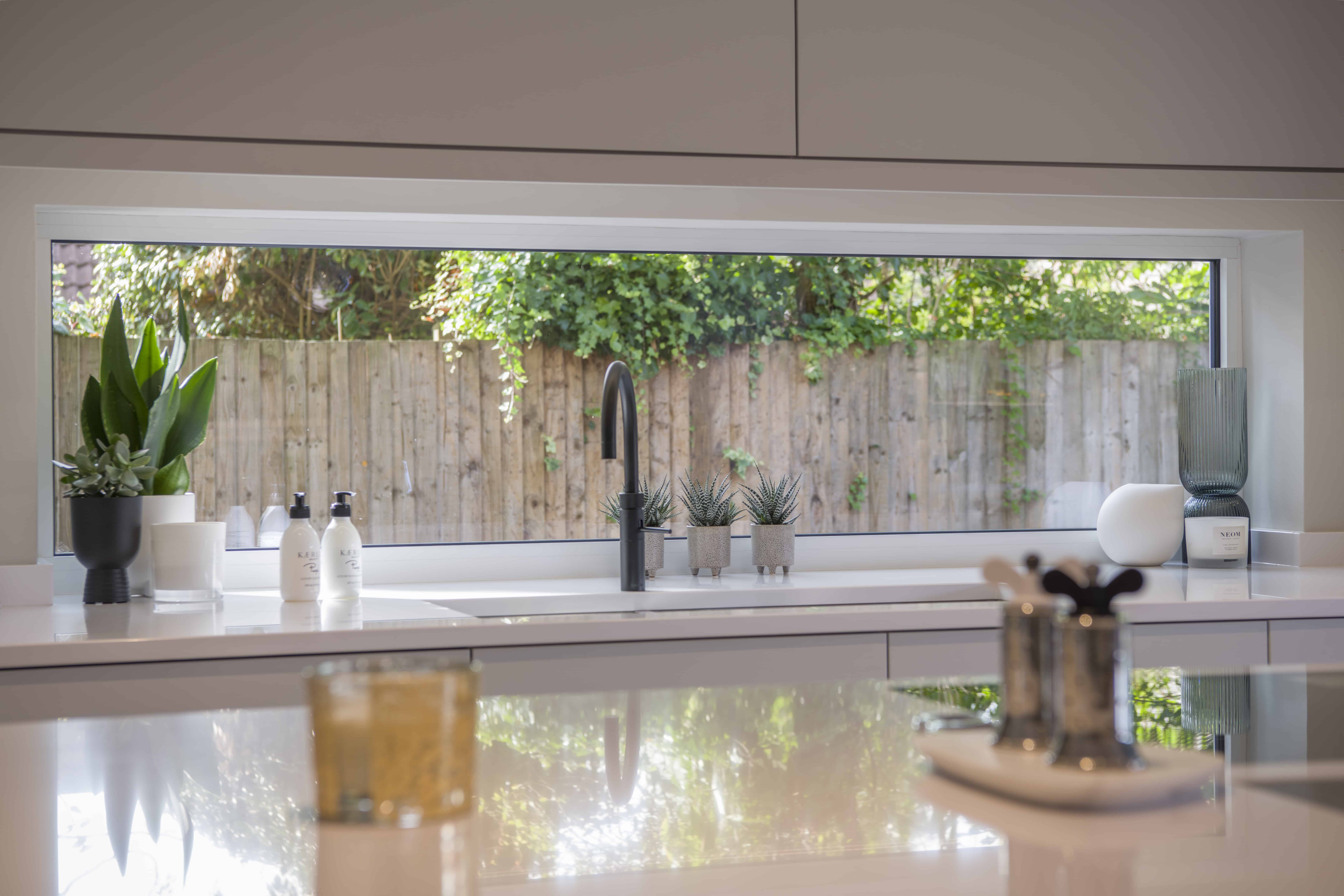 Modern kitchen with a sleek white countertop, potted plants, soap dispensers, and a black faucet in front of a wide rectangular window overlooking sliding doors and greenery outside.