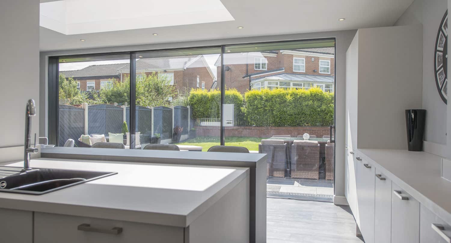 Modern kitchen with white cabinets and an island sink, featuring large floor-to-ceiling sliding doors that open to a sunny backyard with a dining table, chairs, and trimmed hedges.