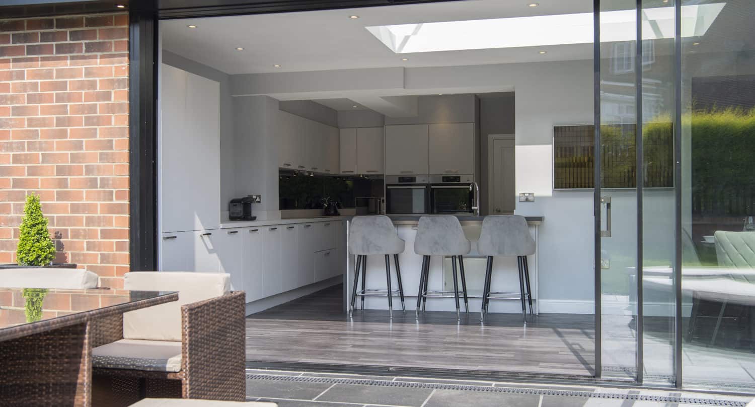 Modern kitchen with white cabinets and a breakfast bar with three gray stools, viewed through open sliding doors from a tiled patio area with outdoor seating. Natural light streams in from a skylight above.