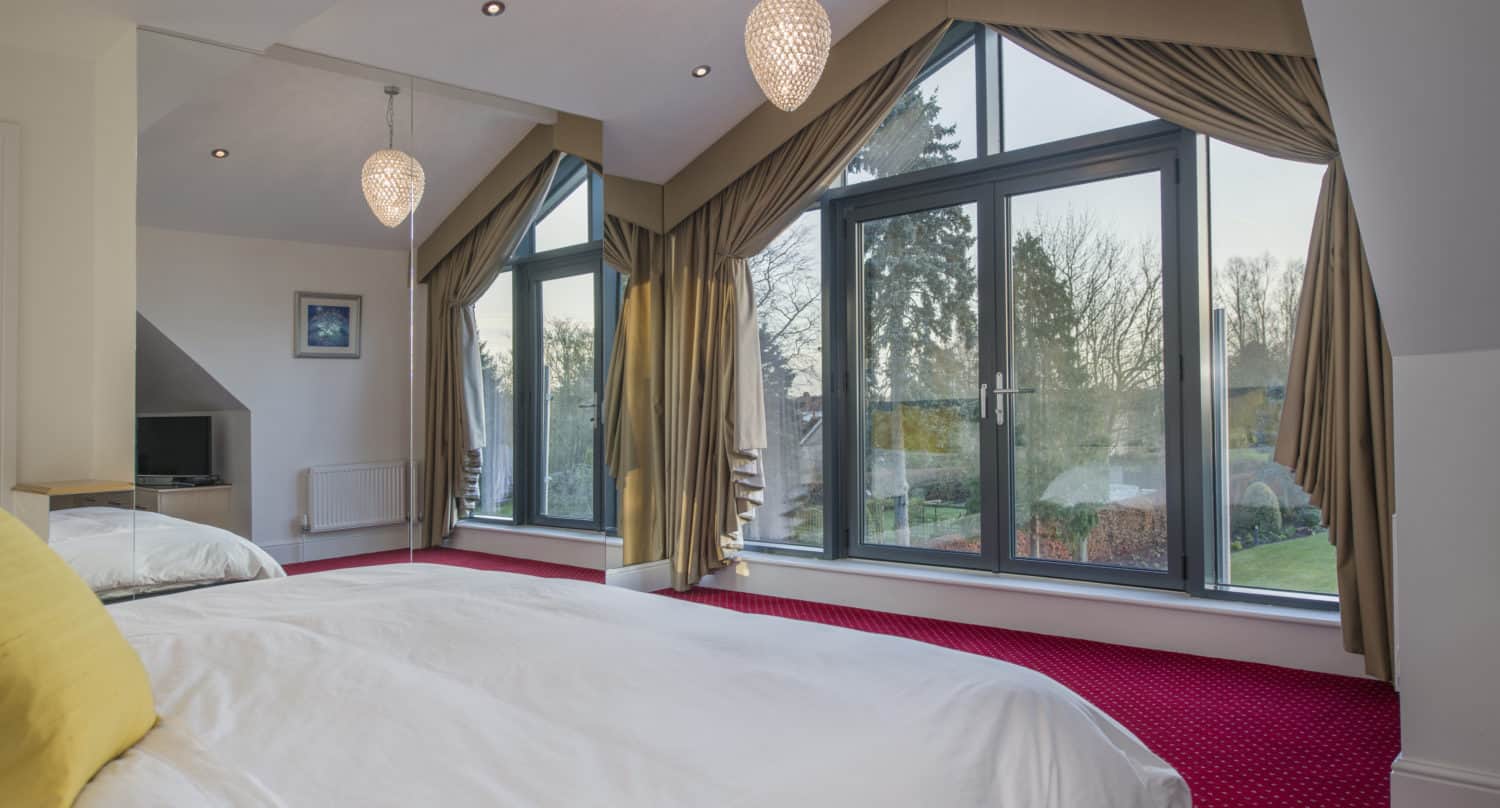 A modern bedroom with sloped ceilings, large floor-to-ceiling windows with gold curtains, a red carpet, white bedding with a yellow pillow, and mirrored walls reflecting the bed and sliding doors.