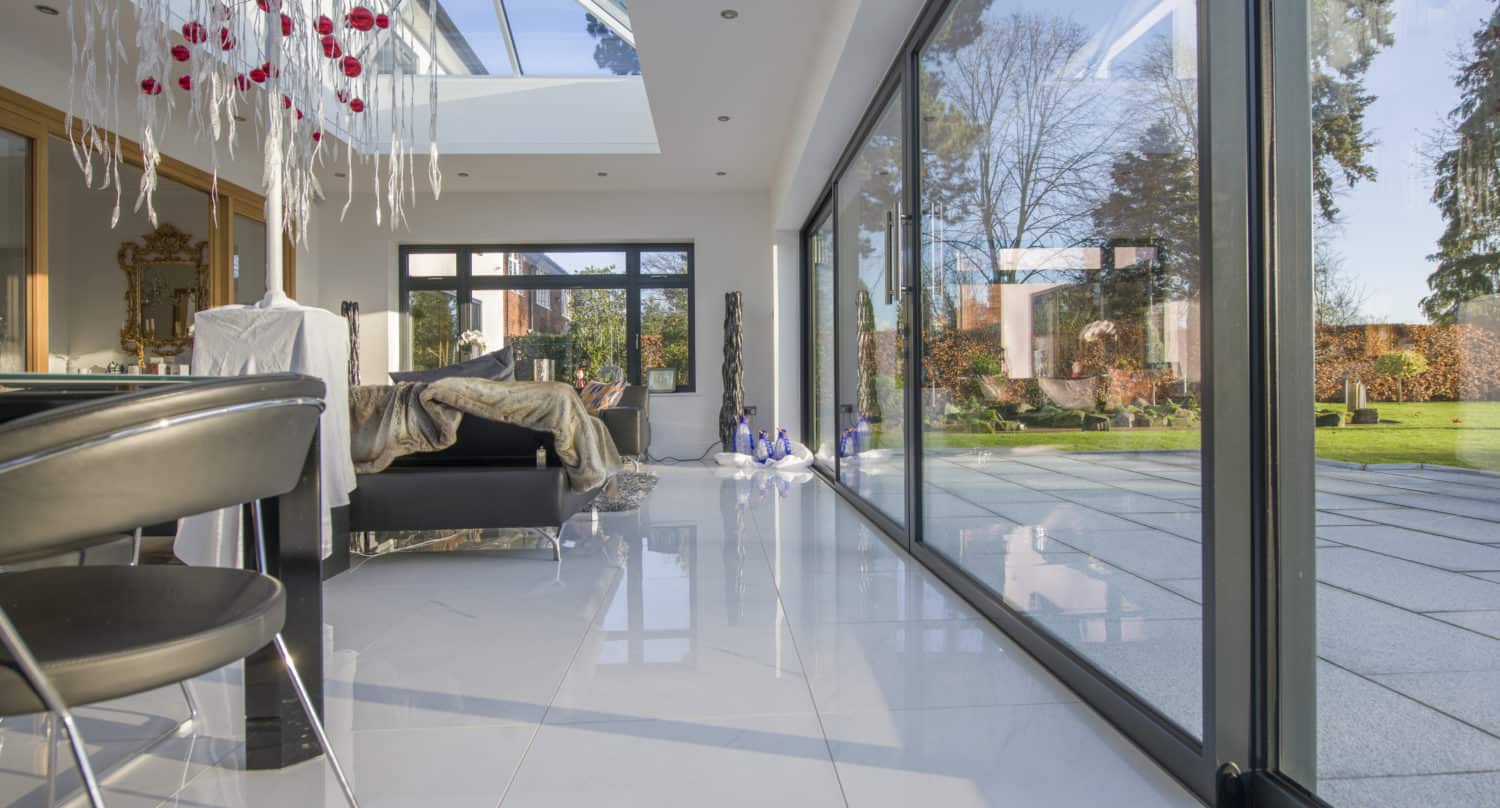 Modern bedroom with large bifold doors opening to a patio and garden, white tile floor, unique white and red chandelier, and skylights letting in natural light.