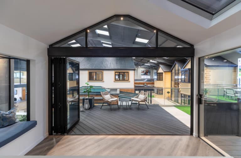 Modern indoor space with large sliding glass doors opening to a balcony with outdoor seating, overlooking a courtyard featuring contemporary buildings and striking glass roofing.