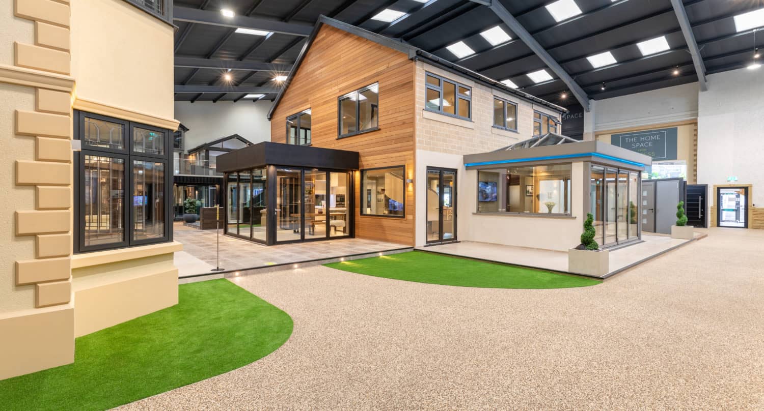 A spacious showroom displays modern house exteriors with large windows, bifold doors, and tan or wood siding, set on a pebble-textured floor with green artificial grass accents under a high, dark ceiling.