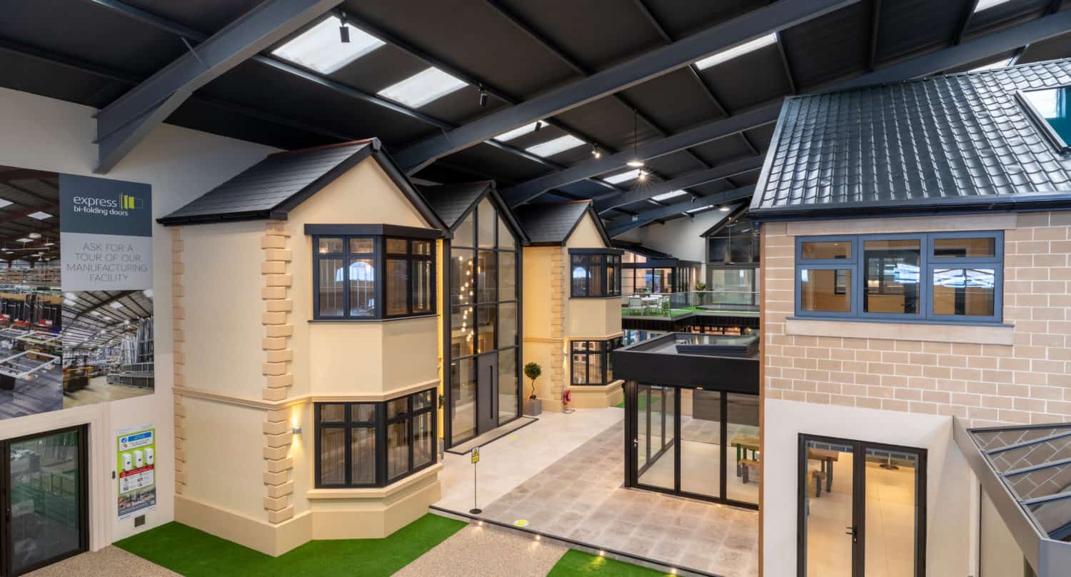 An indoor showroom displays two full-scale model houses with large windows, bifold doors, brick and stone facades, and artificial grass. The space has a high ceiling with skylights and modern lighting.