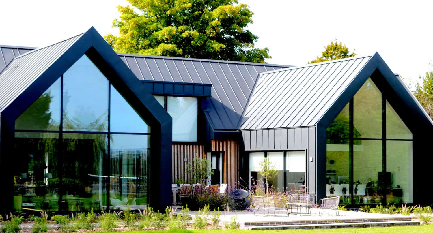 Modern house with dark metal roof, large floor-to-ceiling windows, and bifold doors, surrounded by green lawn, patio seating, and trees in the background on a bright day.