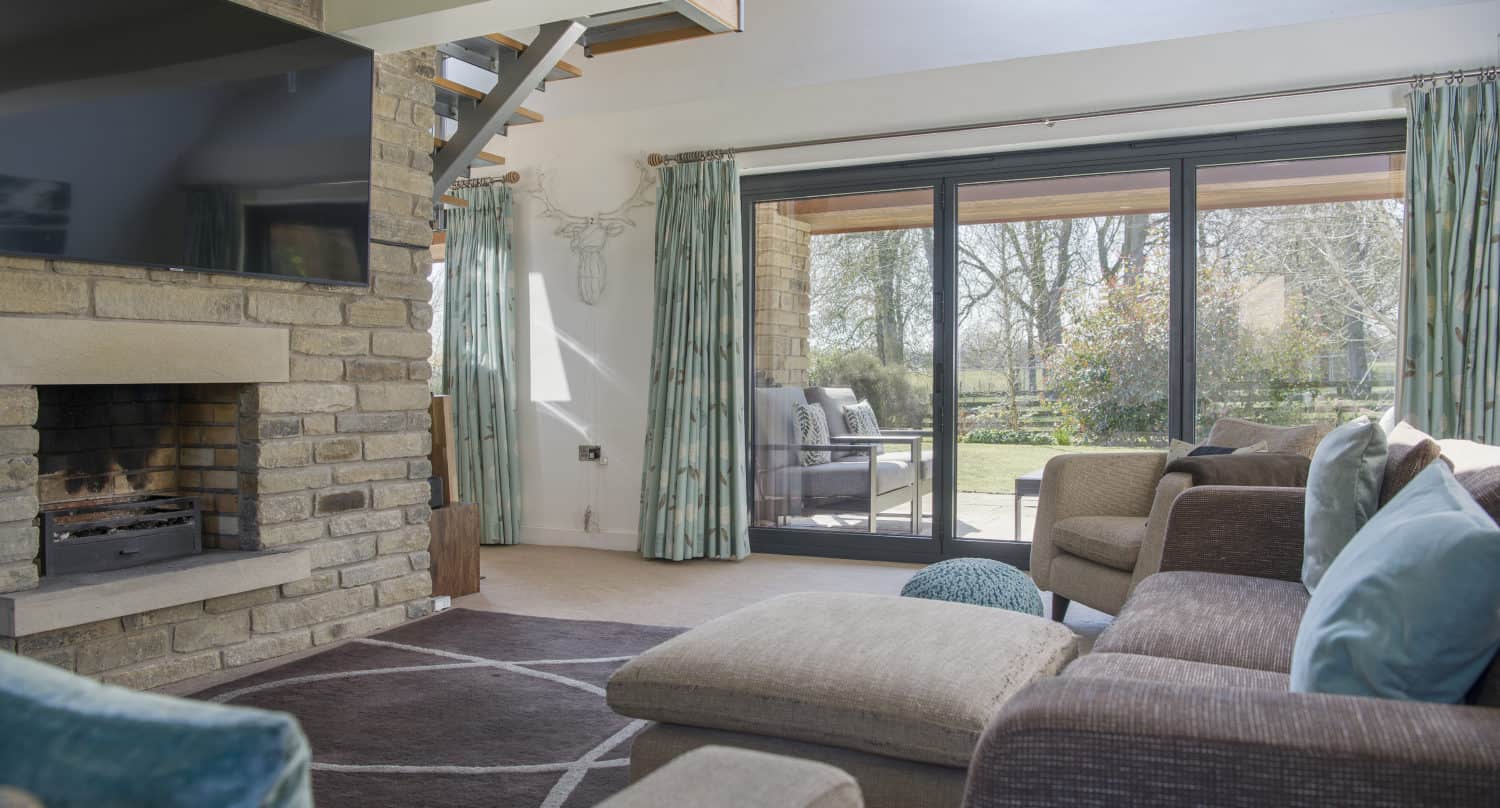 A cozy living room with a stone fireplace, large TV, brown sofas with teal cushions, and sliding doors opening to a sunny patio and garden with trees outside. Light green curtains frame the windows.