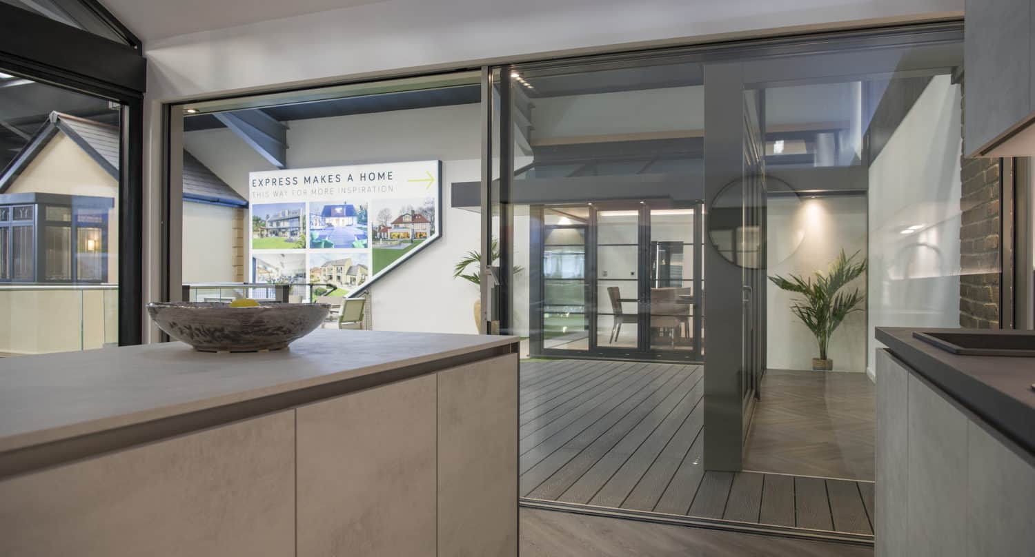 Modern indoor space with large bifold doors opening to a sunlit patio area. Inside, there are light-colored cabinets and a decorative bowl, with leafy plants and signage visible outside.