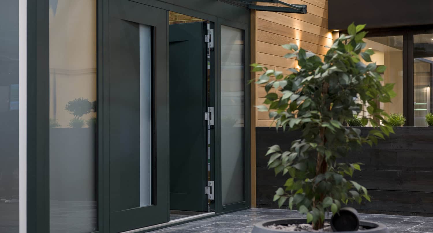 A modern building entrance with glass bifold doors slightly open, featuring an overhang and wood-paneled walls. In the foreground, there is a large potted plant on a tiled floor.
