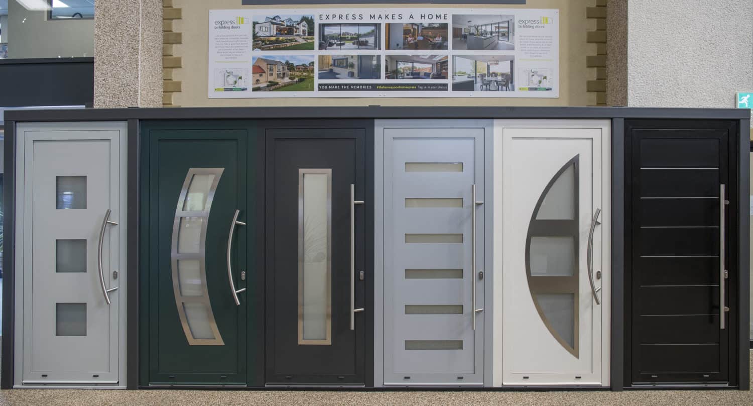 A row of six modern, stylish front doors in different colors and designs is displayed in a showroom, alongside posters highlighting options like sliding doors and bespoke glass solutions above them.