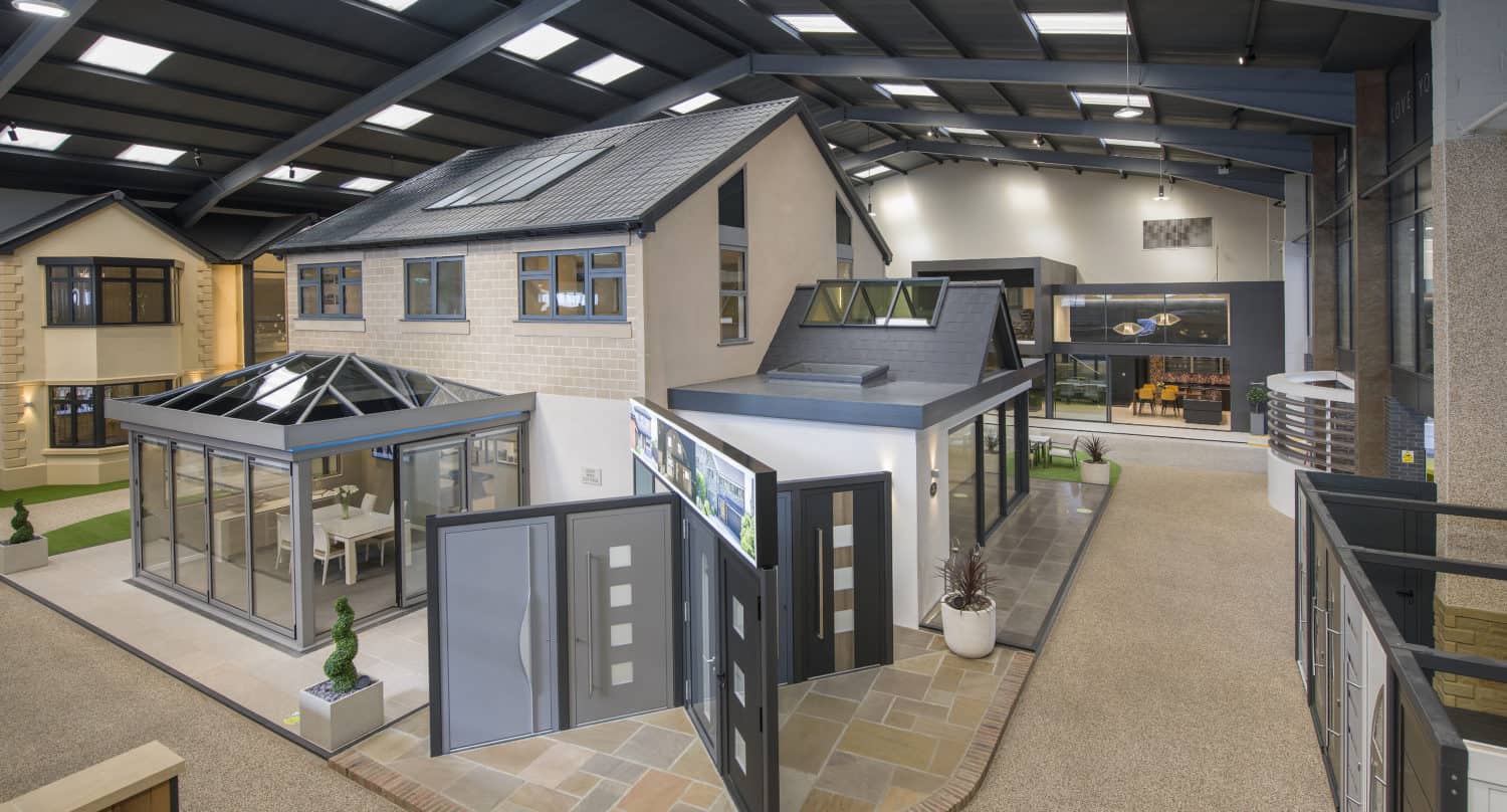 Indoor showroom featuring modern house models with glass doors, front doors, windows, and conservatories under a high ceiling with skylights. The well-lit area showcases various building and home improvement materials.