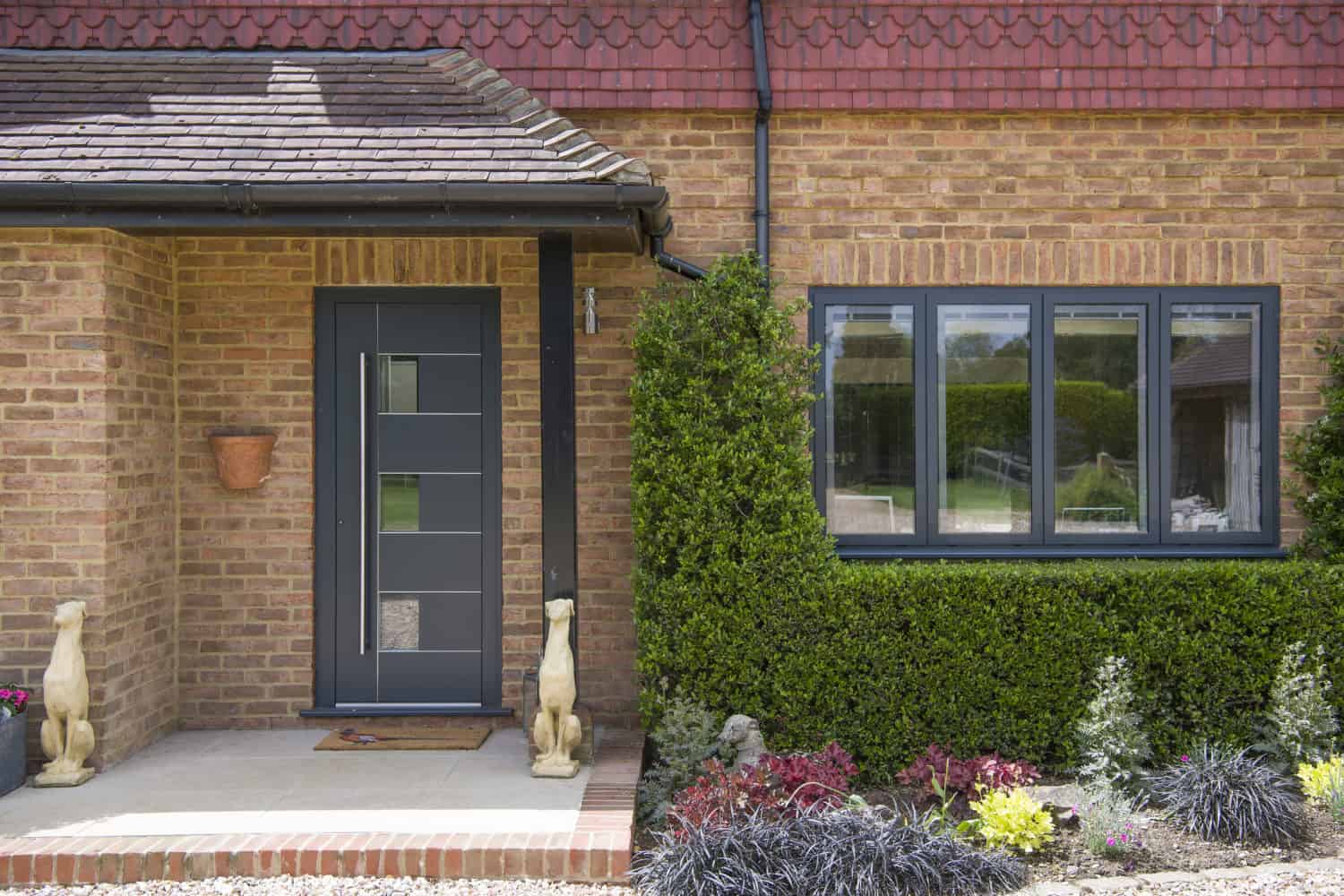 Modern brick house exterior with a dark front door, two decorative dog statues on the porch, a potted plant on the wall, glass roofing above the entryway, and shrubs and colorful plants in the landscaped garden.