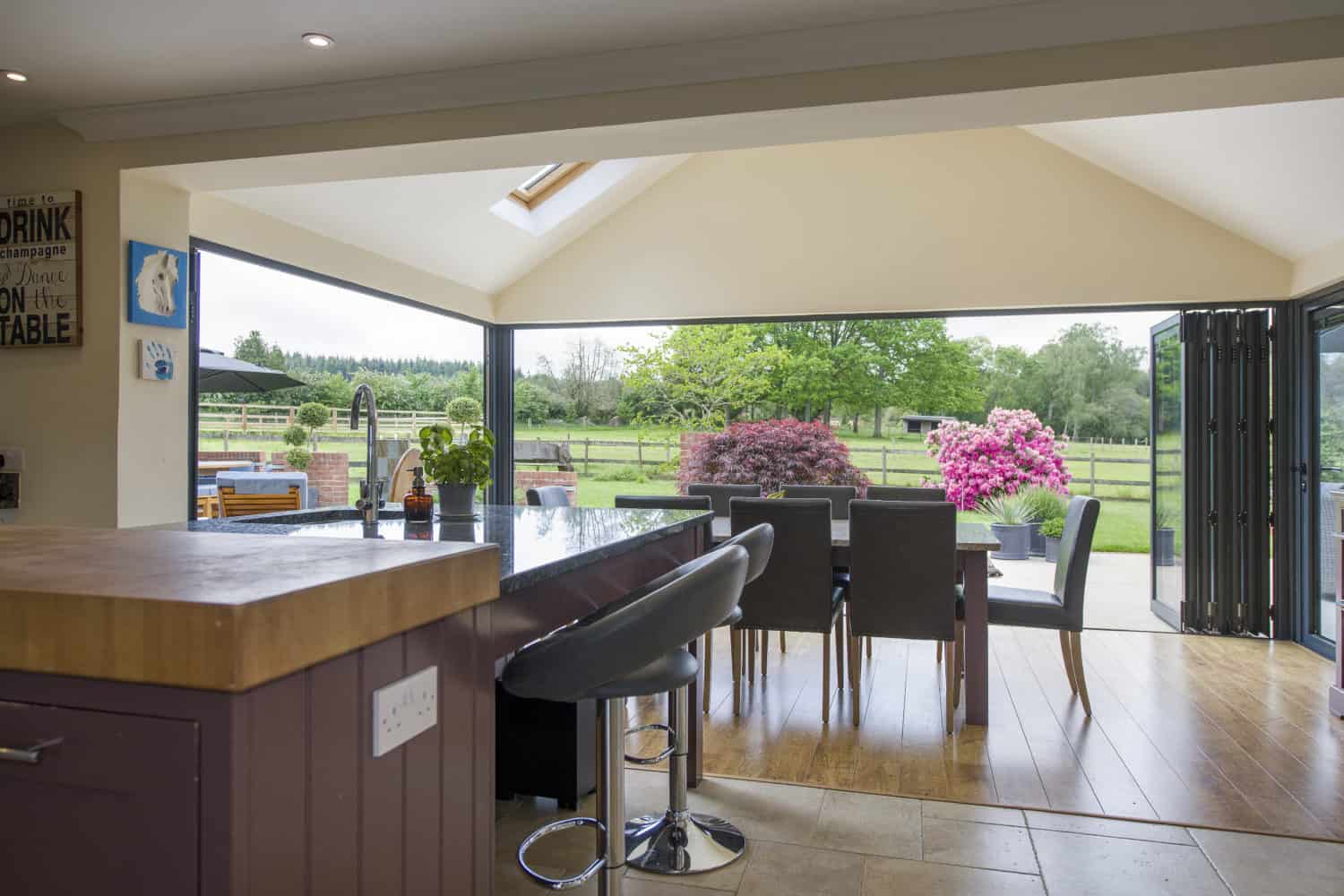 Modern open-plan kitchen and dining area with bifold doors leading to a patio and garden, featuring a large island, barstools, and views of vibrant greenery and pink flowering bushes outside.
