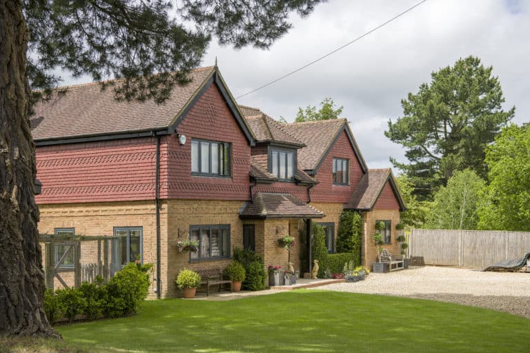 A large brick and red-tiled house with multiple gables, black-framed windows, bifold doors, potted plants, hanging flower baskets, and a well-kept lawn, surrounded by trees and a gravel driveway under a partly cloudy sky.