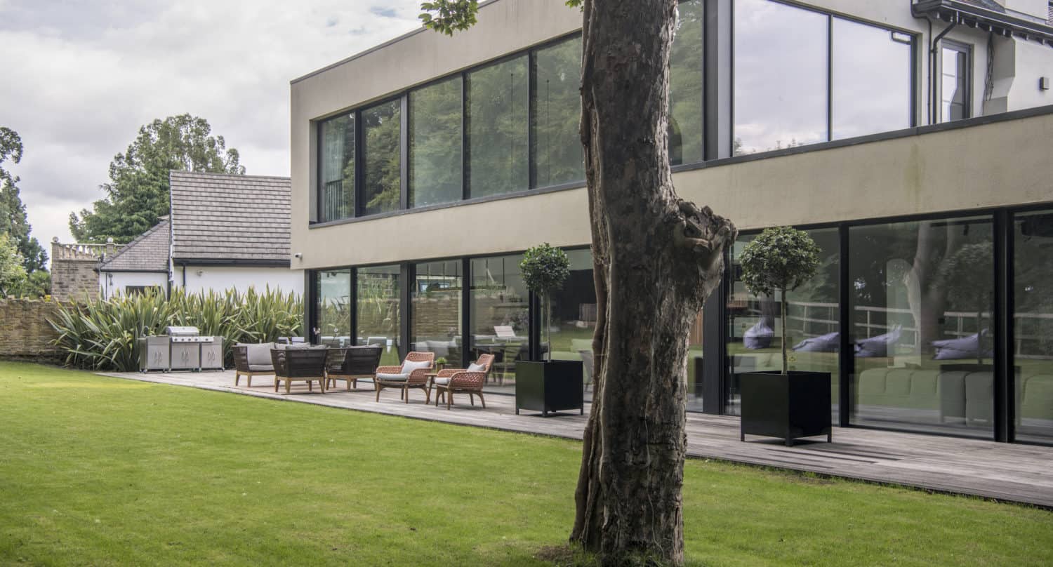 A modern house with large floor-to-ceiling windows and sleek sliding doors, a wooden deck with outdoor seating, and a grassy lawn with a tree in the foreground. Shrubs line the side of the house under a cloudy sky.