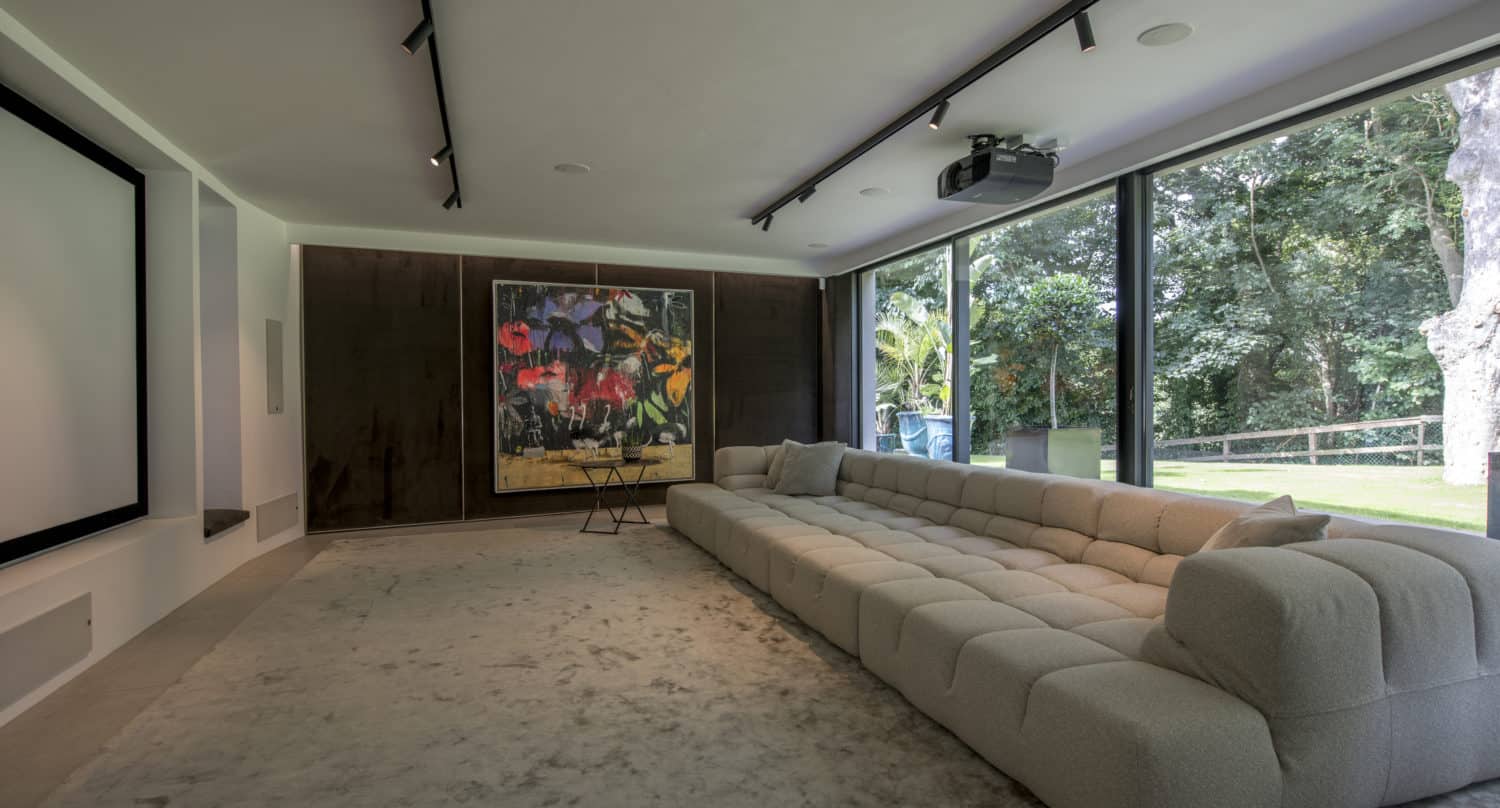 Modern home theater room with a large projector screen, plush beige sectional sofa, abstract painting on the wall, and sliding doors leading to floor-to-ceiling windows overlooking a lush garden.