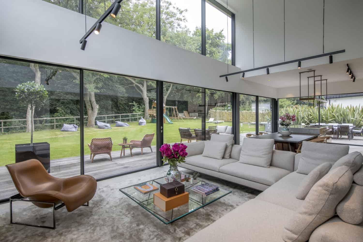 A modern living room with large bespoke glass solutions for floor-to-ceiling windows overlooking a green yard. The space features a light gray sectional sofa, a brown lounge chair, and a glass coffee table with pink flowers and books.