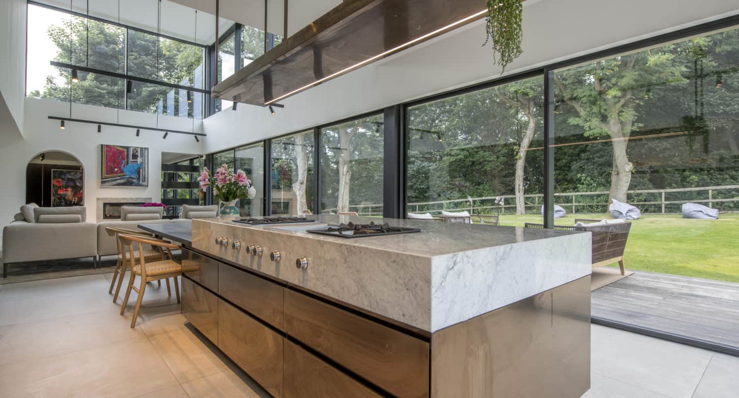 Modern kitchen with a marble island, wooden cabinets, and large floor-to-ceiling windows overlooking a green garden. Open-plan layout with bespoke glass solutions, dining area, living space, and contemporary decor.