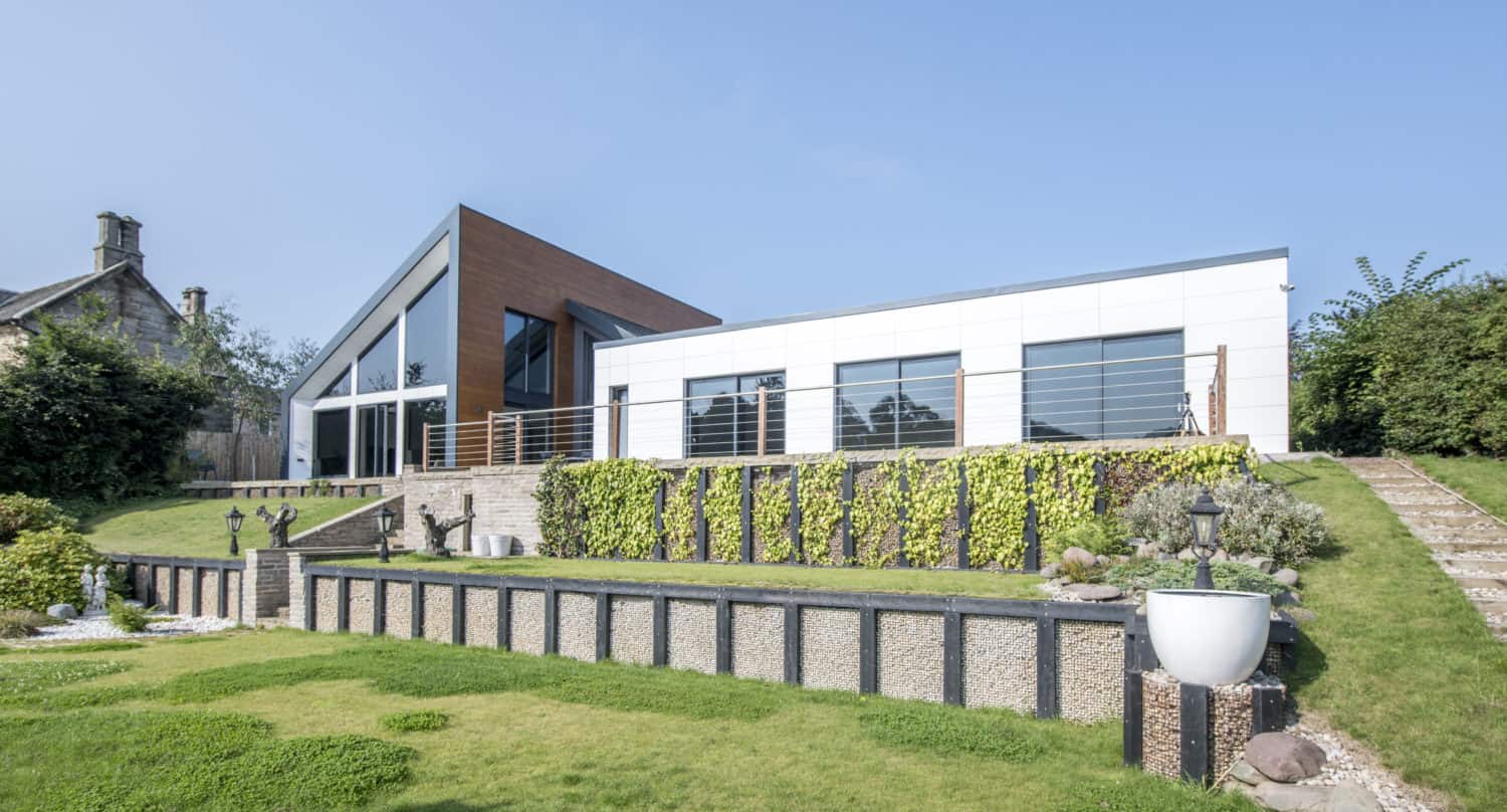 A modern house with large windows, bifold doors, and an angular roof sits on a landscaped lawn with terraced stone walls, green grass, and decorative plants under a clear blue sky.