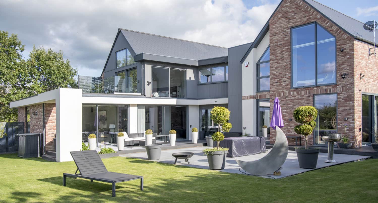 Modern house with large glass windows and brick accents, featuring bifold doors that open to a landscaped backyard with patio furniture, potted plants, and a lounge chair on a grassy lawn under a partly cloudy sky.