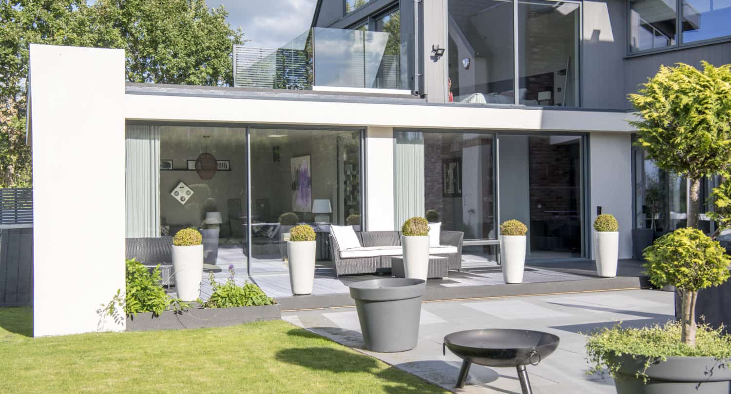 Modern house with large bifold doors, a patio with outdoor seating, tall white planters, a fire pit, and a neatly manicured lawn, surrounded by greenery and trees on a sunny day.