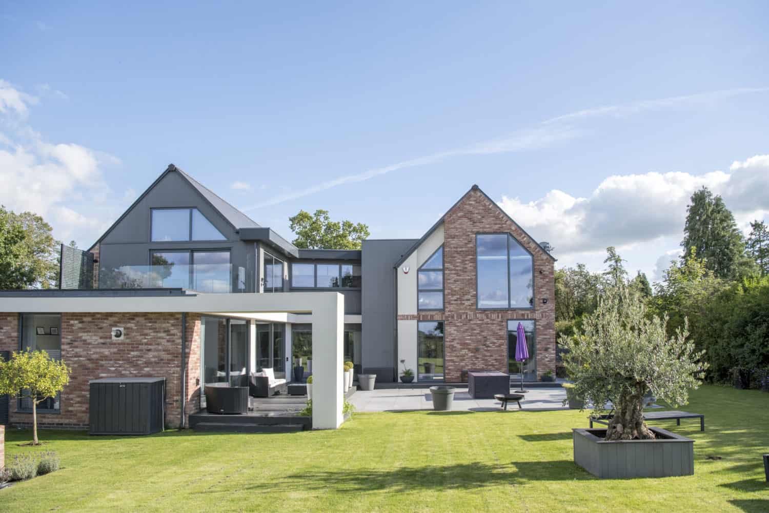 A modern two-story house with large glass windows, bifold doors, a brick and gray exterior, spacious lawn, outdoor seating, potted plants, and trees under a blue sky with scattered clouds.