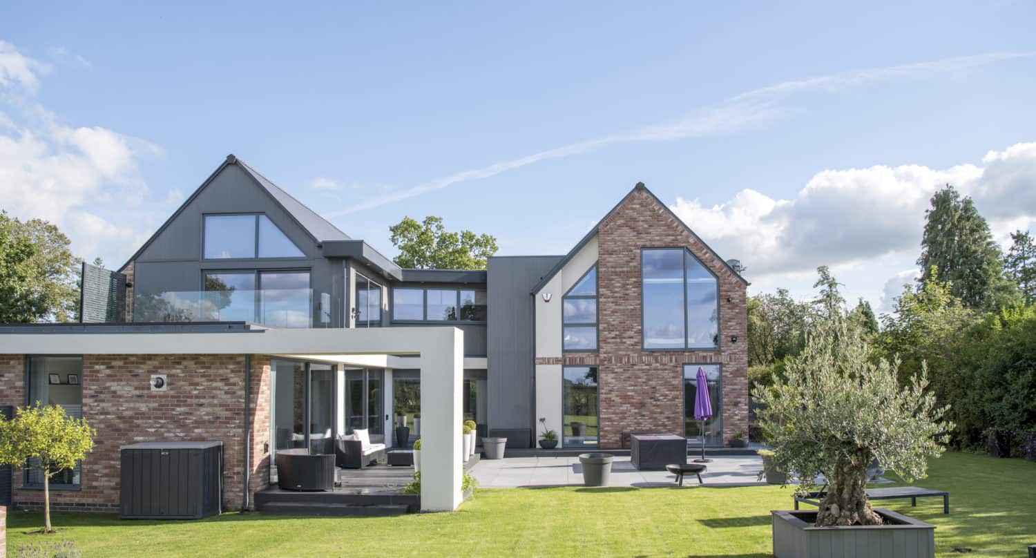 A modern two-story house with large glass windows, bifold doors, a brick and gray exterior, spacious lawn, outdoor seating, potted plants, and trees under a blue sky with scattered clouds.