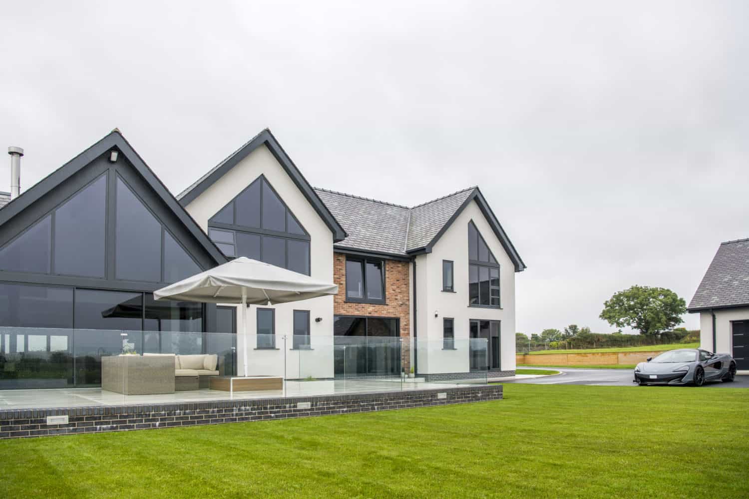 Modern two-story house with large glass windows, stylish sliding doors, a mix of white and brick exterior, and a spacious patio with outdoor seating and an umbrella. A sleek black car is parked on the driveway next to a well-kept lawn.