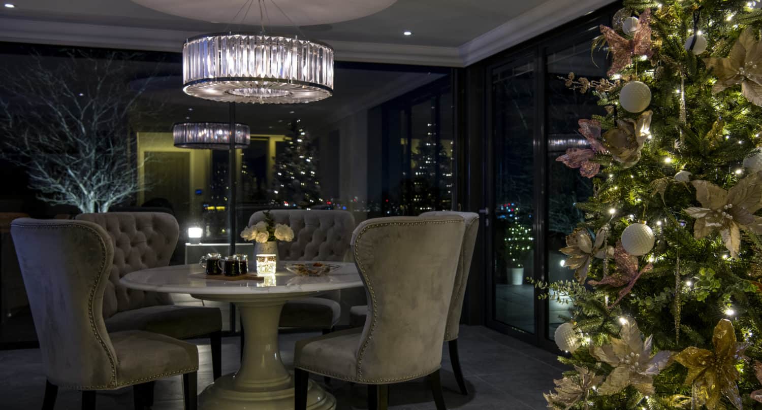 A cozy dining area with a round table and four tufted chairs is softly lit by a crystal chandelier. A decorated Christmas tree with lights and ornaments stands in the foreground by large windows featuring bespoke glass solutions at night.