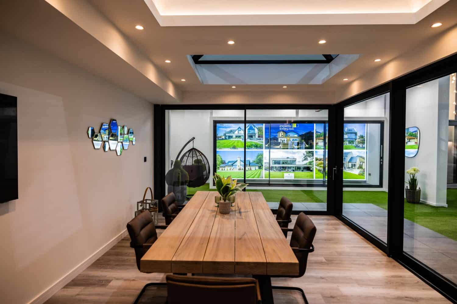 Modern conference room with a long wooden table, brown chairs, and a plant centerpiece. Glass walls display property ads, while glass roofing and ceiling lights brighten the space. A hanging chair sits in the corner.