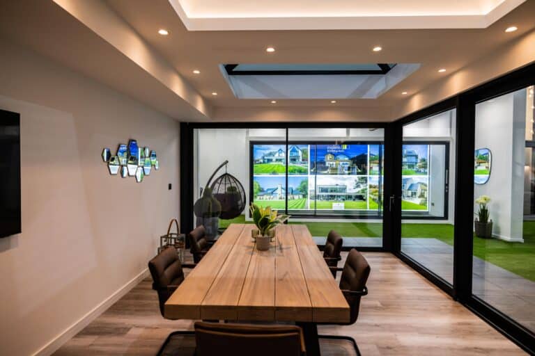 Modern conference room with a long wooden table, brown chairs, and a plant centerpiece. Glass walls display property ads, while glass roofing and ceiling lights brighten the space. A hanging chair sits in the corner.
