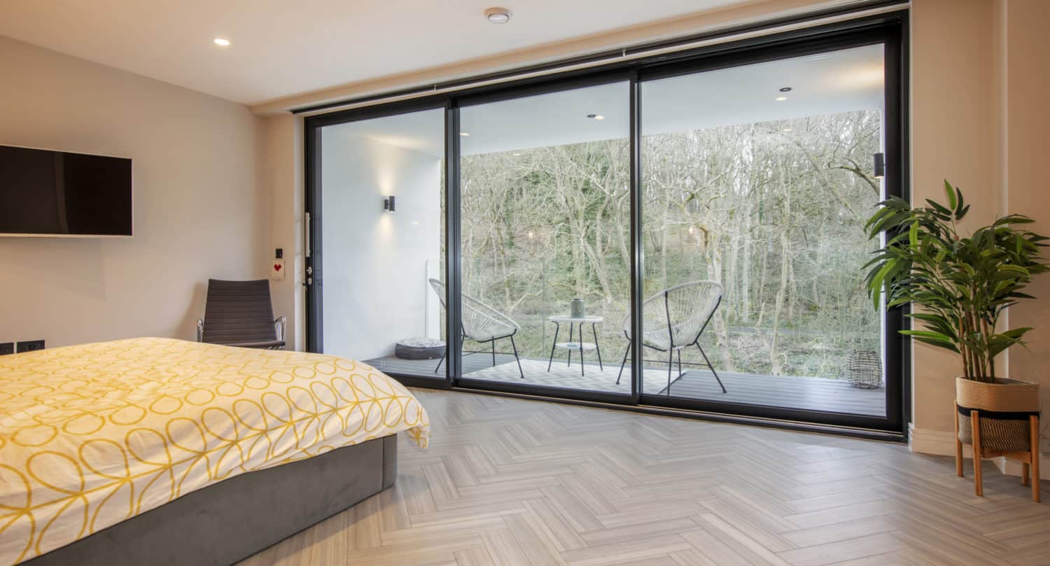Modern bedroom with a patterned yellow and white bed, large bespoke glass solutions for sliding doors leading to a balcony with two chairs and a table, overlooking trees. Light wood herringbone floor, indoor plant, and wall-mounted TV.