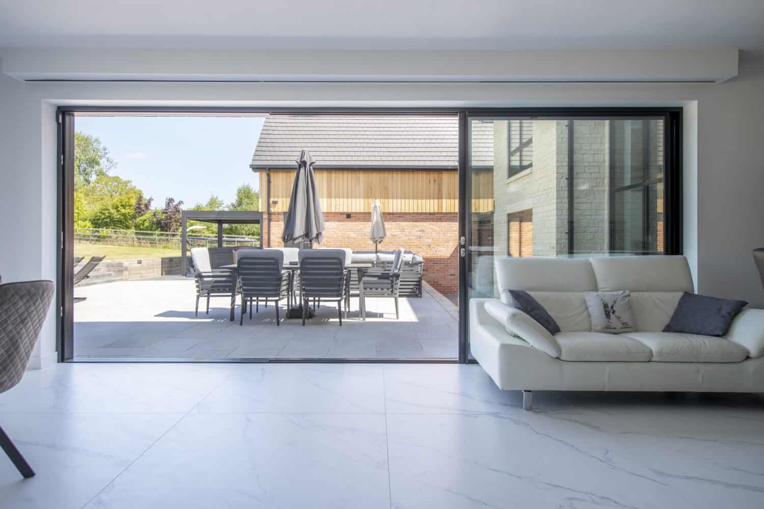 Modern living room with a white sofa and stylish bifold doors opening to a patio with outdoor dining furniture, umbrellas, and a view of a wooden building and greenery outside.