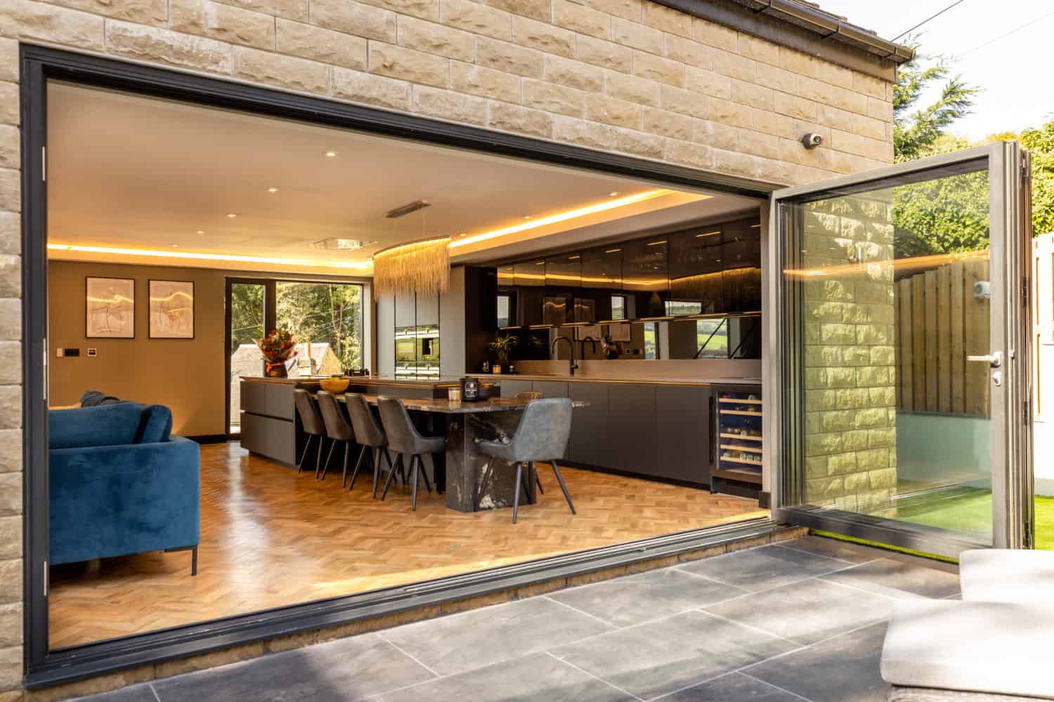 Modern open-plan kitchen and dining area with parquet flooring, visible through large bifold doors opening to a patio. Sleek cabinets, dining table with chairs, and a blue sofa are inside, all enhanced by bespoke glass solutions.