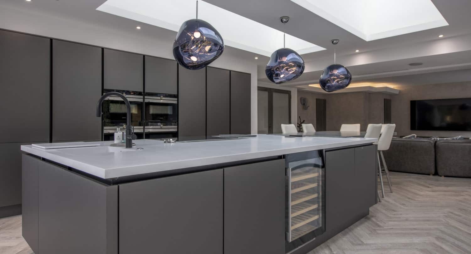 Modern kitchen with a large gray island, integrated wine fridge, sleek cabinetry, and three black pendant lights. Skylights and glass roofing flood the space with natural light. White bar stools and part of a living area are visible in the background.