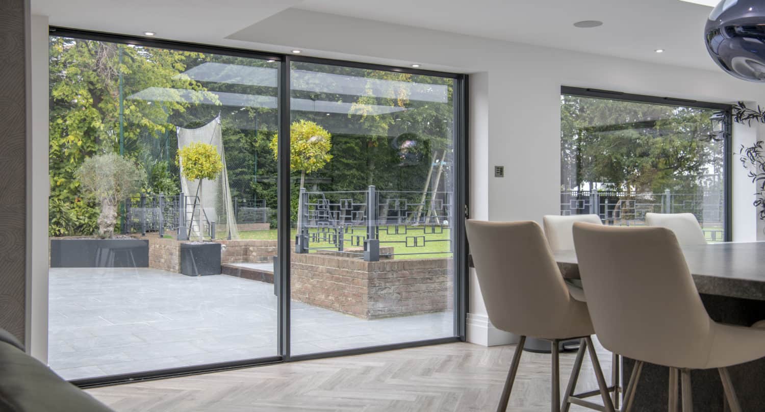 Modern dining area with tall white chairs beside sleek sliding doors, offering a view of a green garden with brick planters, outdoor seating, and trees. Light wood flooring enhances the indoor space.