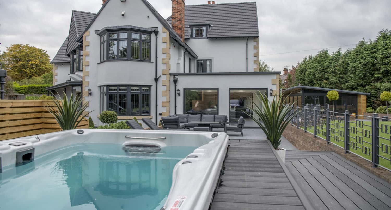 Modern white house with black trim, large windows, and a spacious patio area with outdoor furniture. Sliding doors open to a deck with a hot tub in the foreground, surrounded by greenery and a fenced yard.