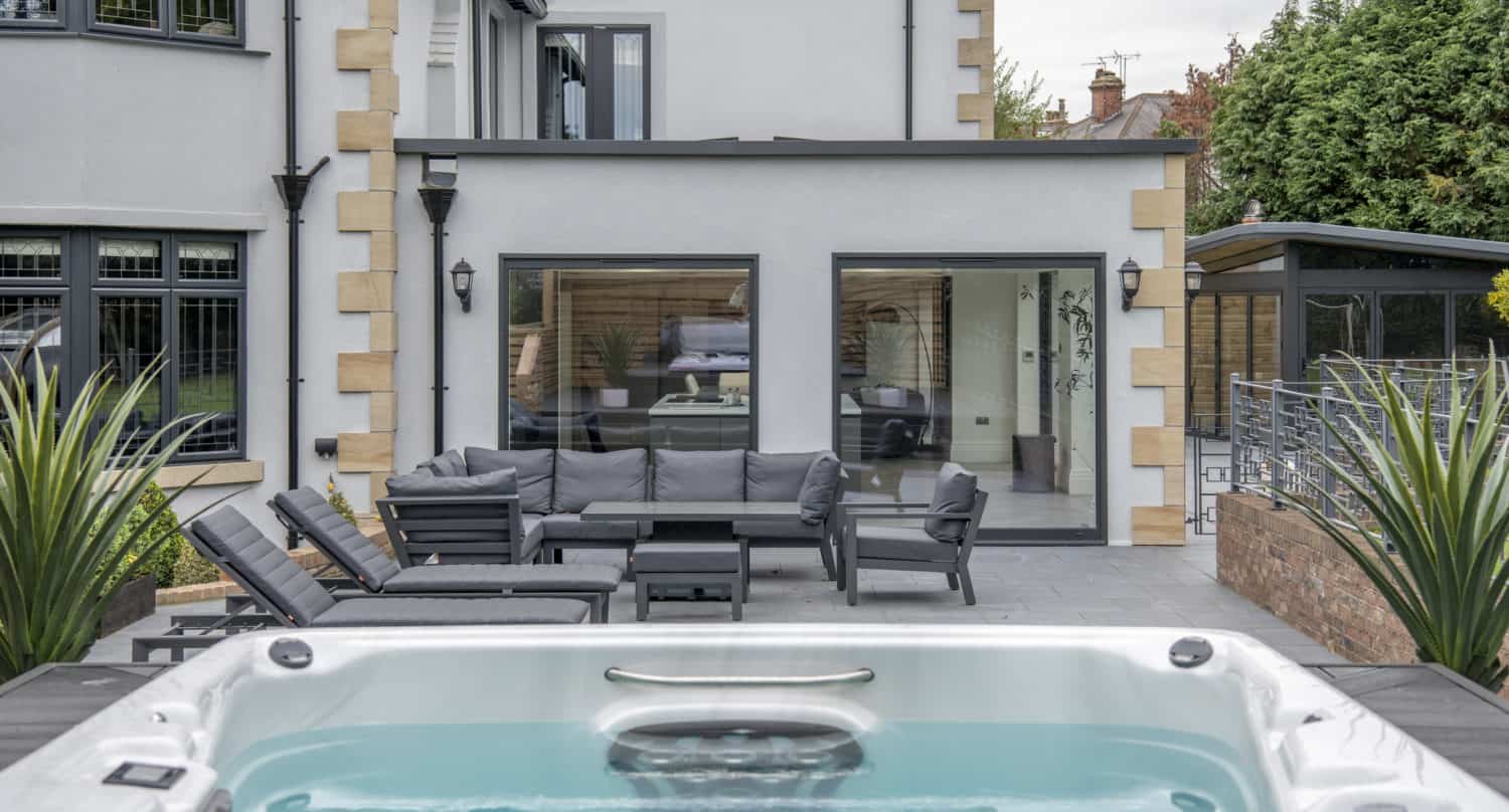 A modern backyard featuring a hot tub in the foreground, an outdoor seating area with gray cushions, and a contemporary house with large windows and sleek sliding doors in the background.