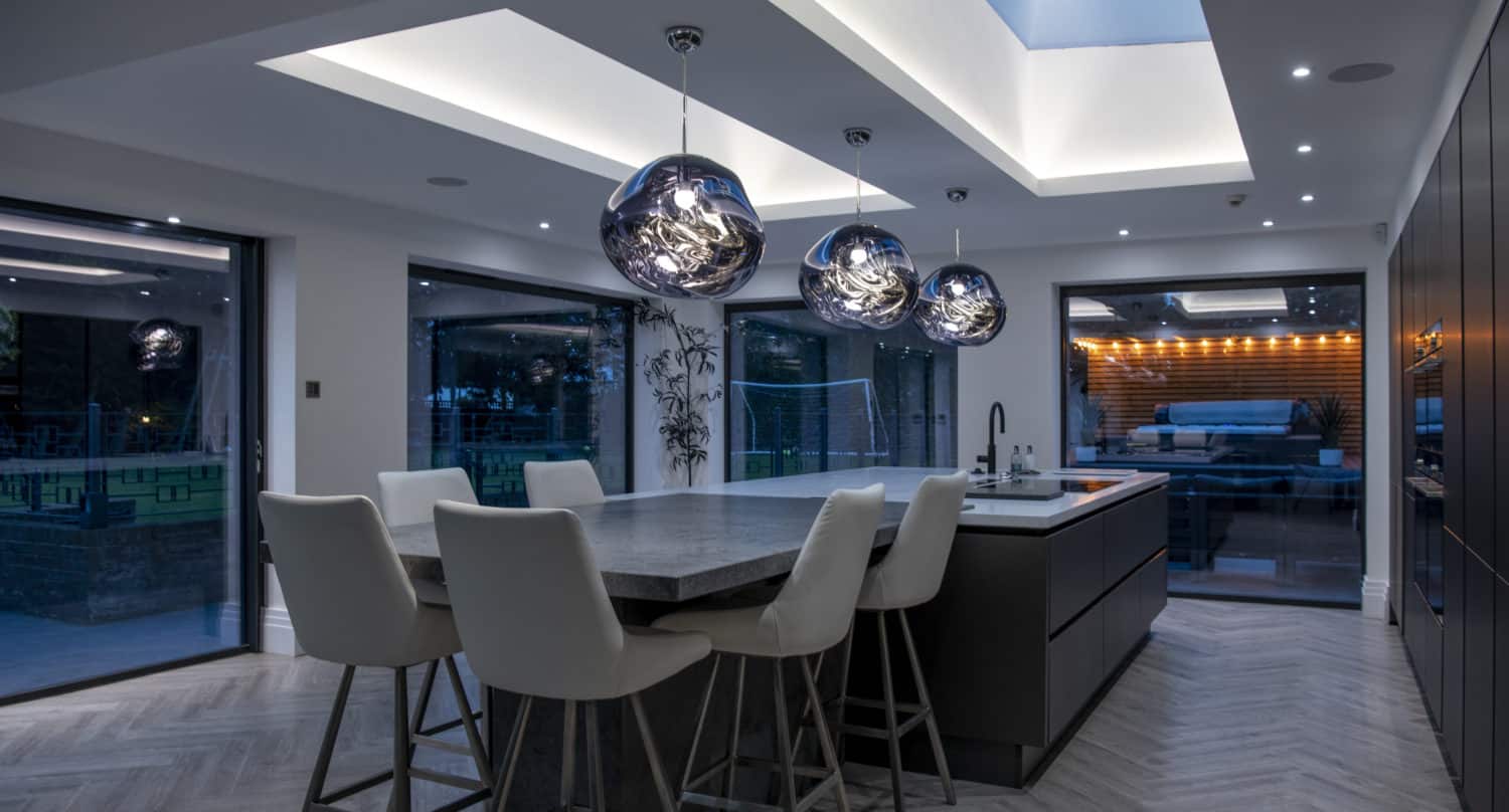 Modern kitchen with a large island, white high-backed chairs, and three artistic pendant lights. Skylights and bespoke glass solutions flood the space with natural light, with an outdoor seating area visible through bifold doors.