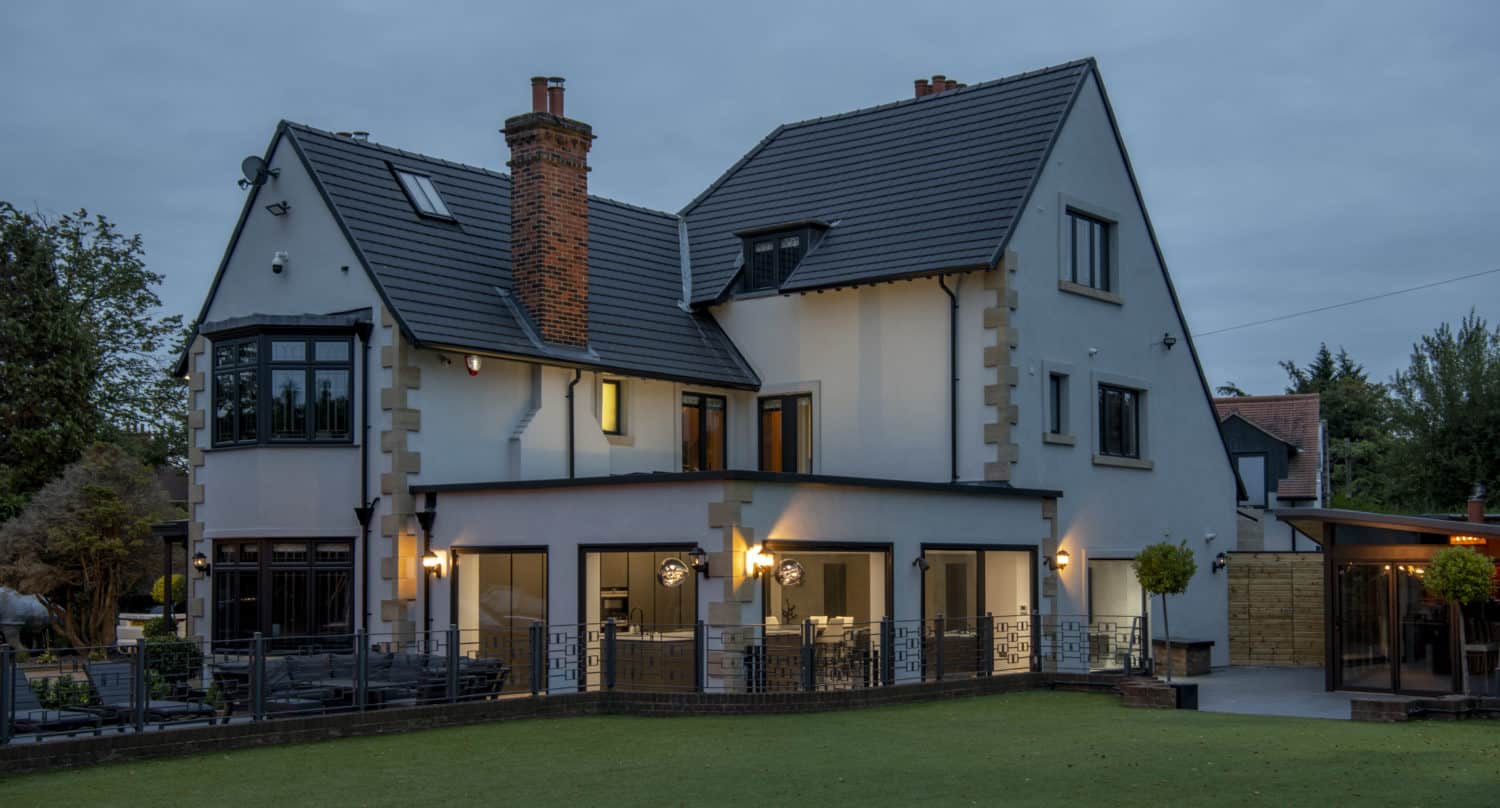 A large, modern two-story white house with black trim, lit warmly at dusk. The home features multiple large windows and sliding doors, a fenced patio area, and a well-kept lawn. Trees and outdoor lights surround the property.