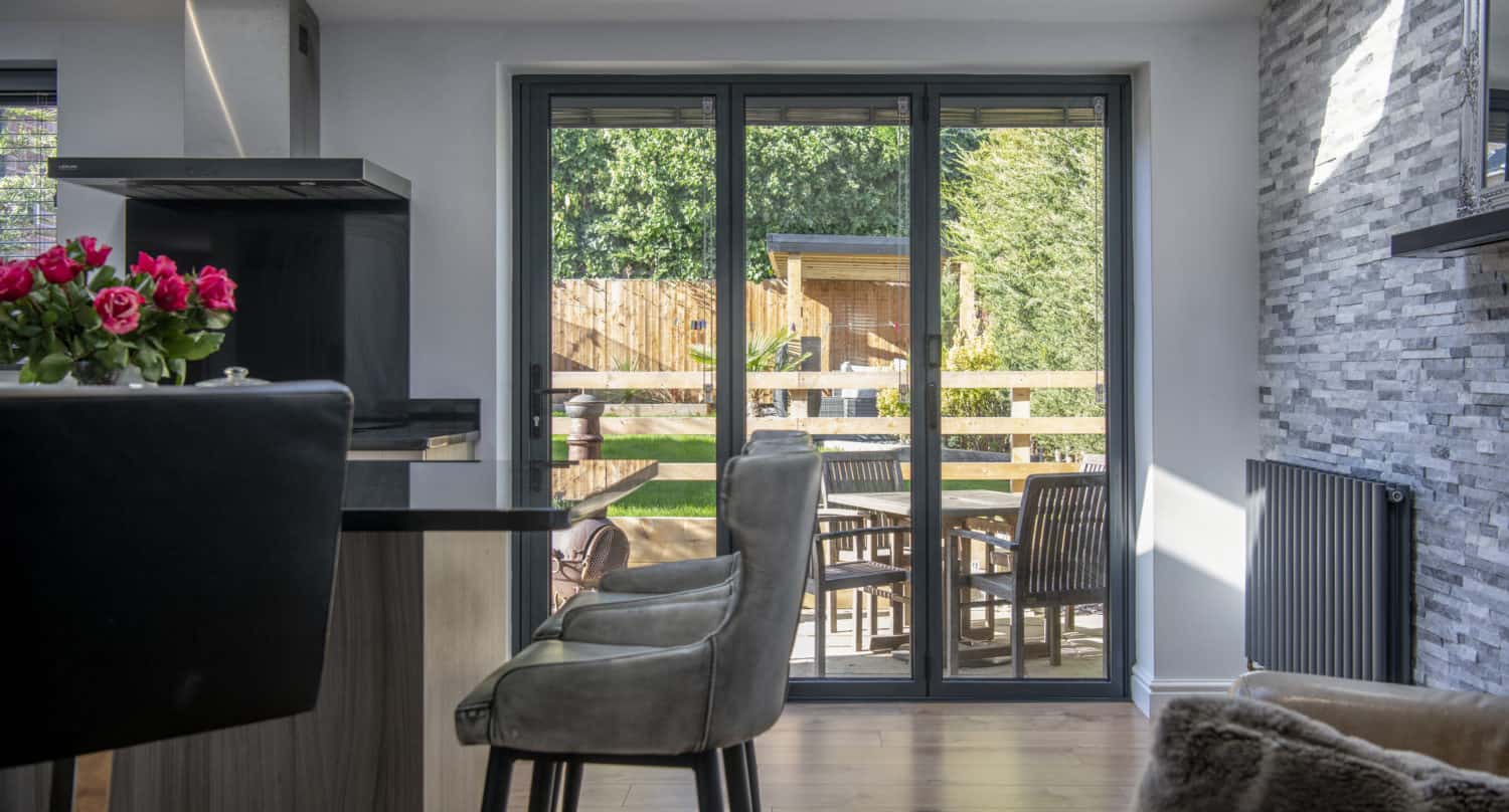 Modern kitchen interior with a breakfast bar and chairs, looking out through bespoke glass solutions and large sliding doors onto a patio with outdoor furniture and a lush green garden beyond.