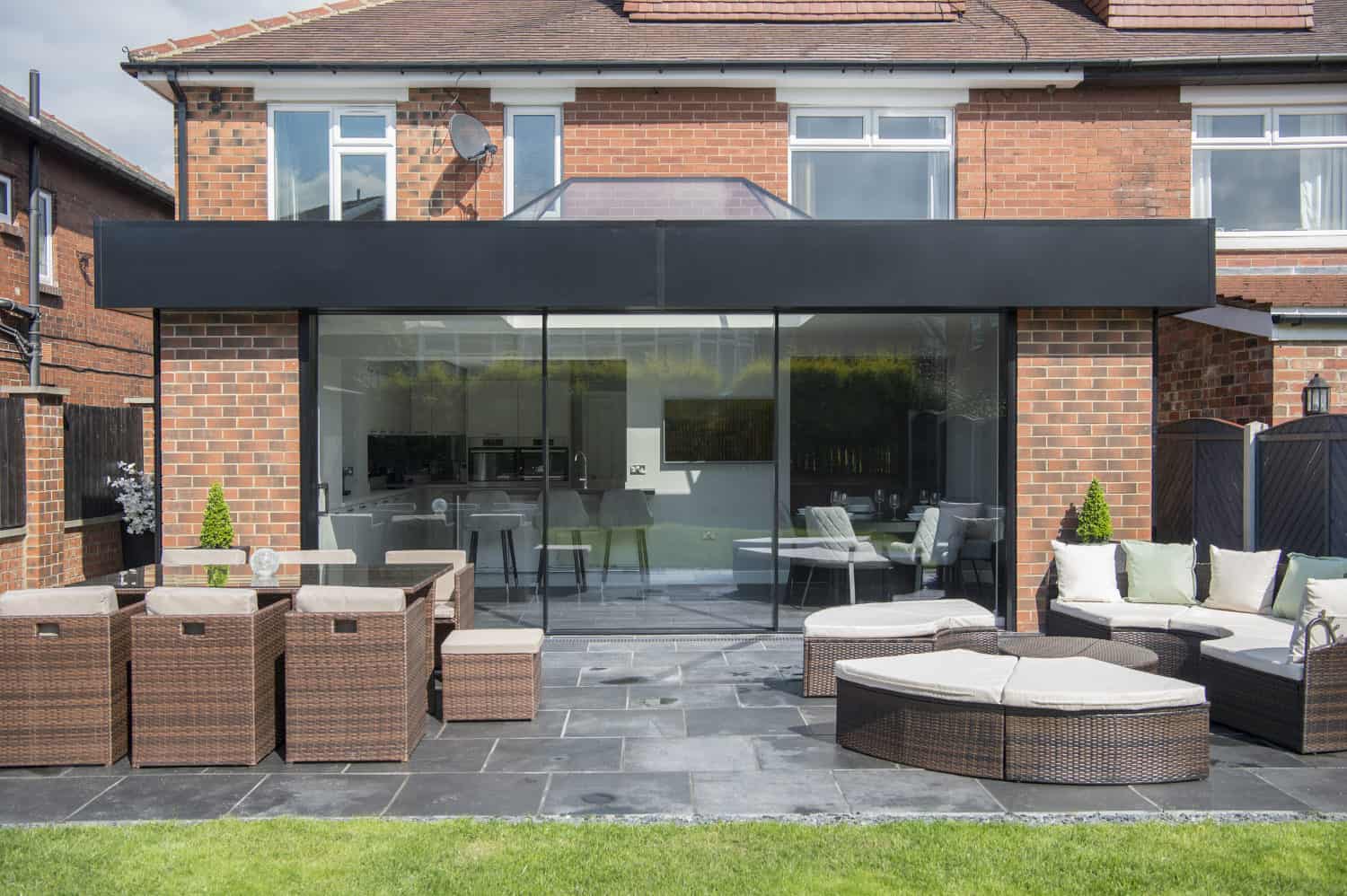 Modern brick house with large glass sliding doors opening to a patio with wicker outdoor furniture, including tables, chairs, and sofas on dark stone tiles, overlooking a green lawn and shaded by partial glass roofing.
