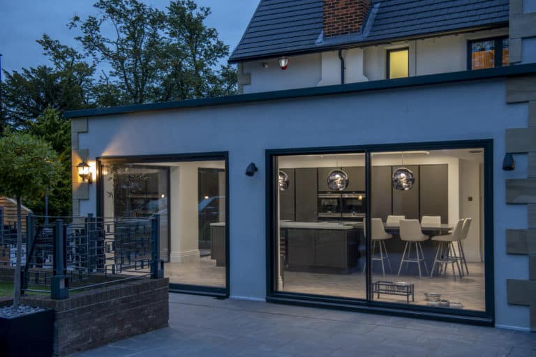 Modern house exterior at dusk featuring expansive bifold doors that reveal a contemporary kitchen and dining area with bar stools; outdoor lights warmly illuminate the inviting patio space.