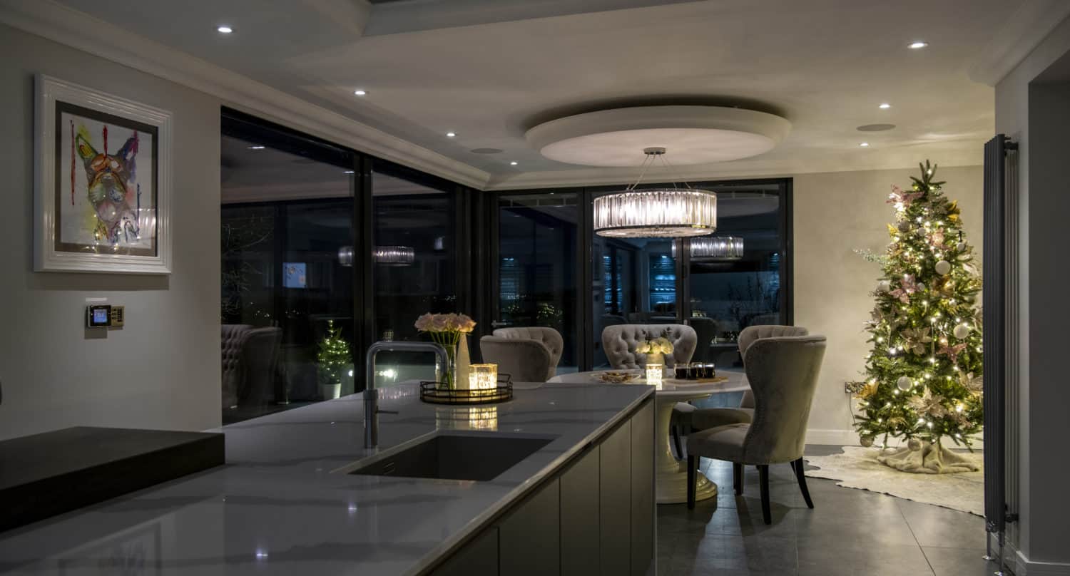 Modern kitchen and dining area decorated for Christmas, featuring a lit Christmas tree, elegant dining chairs, a round table, contemporary lighting, and large windows with bespoke glass solutions revealing a nighttime city view.