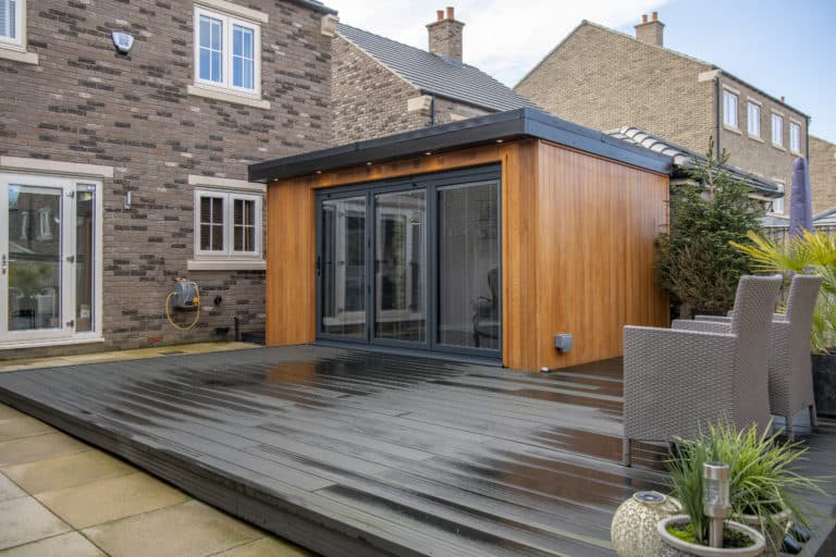 Modern backyard with a wooden garden room featuring large sliding doors, a dark deck, outdoor rattan chairs, potted plants, and the exterior of a brick house in the background.