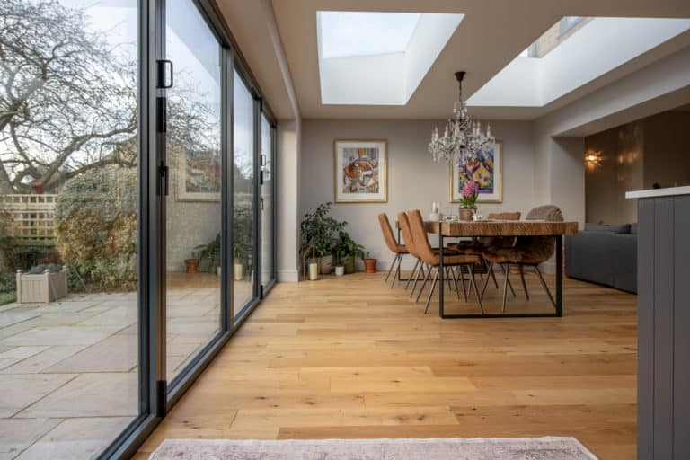A modern dining area with glass roofing, sliding doors, a wooden table with tan chairs, indoor plants, and colorful artwork on light walls, overlooking a patio and garden.